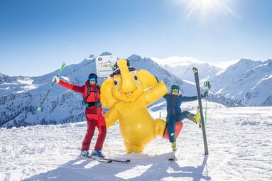 Two skiers pose beside a yellow elephant figure in Hochkrimml, with snow-covered mountains of Zillertal Arena behind them. | © Zillertal Arena