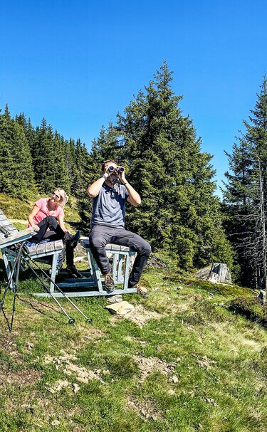 Familie bei einer Wanderpause auf einer Bergwiese mit Fernglas und Wanderstöcken, im Hintergrund Wald und Berge. | © TVB Piesendorf Niedernsill, Foto Harry Liebmann 