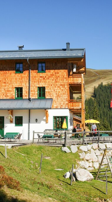 Pinzgauer Hütte with terrace and play area on a mountainside, surrounded by meadows and forest. | © TVB Piesendorf Niedernsill, Foto Harry Liebmann