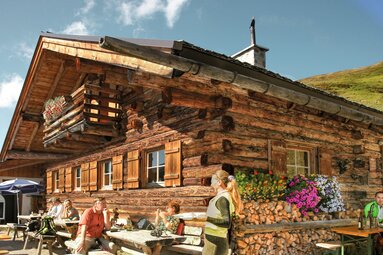 Hochsonnbergalm in Piesendorf mit Gästen an Tischen vor der Holzhütte, Blumenschmuck und gestapeltem Brennholz. | © TVB Piesendorf Niedernsill