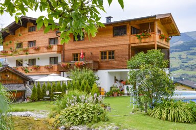 Pension Jausenstation Burgeck with wooden balconies, garden and a round pool in front of green mountain slopes. | © Pension - Jausenstation Burgeck