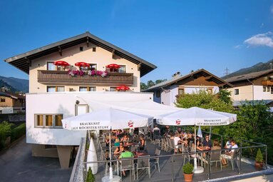 Zur Pinzgauerin with a large terrace, guests under sun umbrellas and a balcony decorated with flowers. | © Gasthof Zur Pinzgauerin