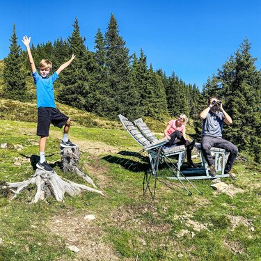 Familie bei einer Wanderpause auf einer Bergwiese mit Fernglas und Wanderstöcken, im Hintergrund Wald und Berge. | © TVB Piesendorf Niedernsill, Foto Harry Liebmann 