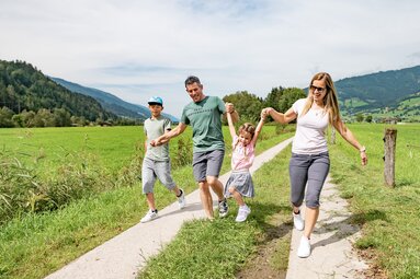 Familie mit zwei Kindern geht auf einem Weg durch Wiesen; die Eltern halten das jüngere Kind beim Springen an den Händen. | © TVB Piesendorf Niedernsill, Foto Lichtfarben