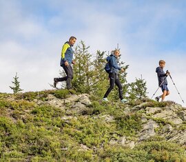 Zwei Erwachsene und ein Junge wandern über einen felsigen Berghang. Der Junge geht mit Wanderstöcken voraus. | © Lichtfarben
