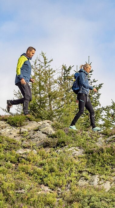 Zwei Erwachsene und ein Junge wandern über einen felsigen Berghang. Der Junge geht mit Wanderstöcken voraus. | © Lichtfarben