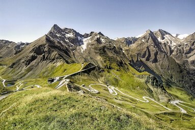 View from Edelweiß-Spitze across mountain peaks with snowfields and a road with several hairpin bends. | © Großglockner.at