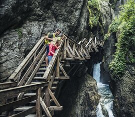 Eine Familie steht auf einem Holzsteg in der Sigmund-Thun-Klamm in Kaprun und blickt auf den Wasserfall. | © Zell am See-Kaprun Tourismus