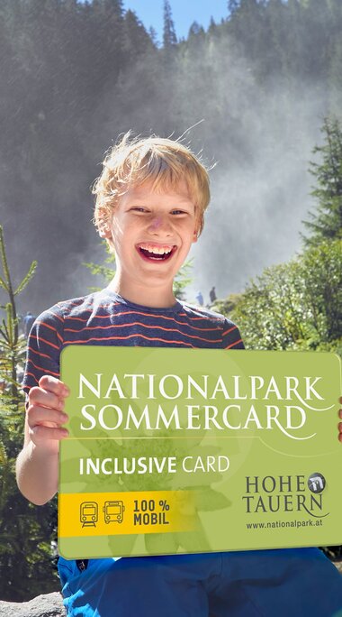 Child holding a Nationalpark Sommercard in front of the Krimml Waterfalls, surrounded by rocks and forest. | © Ferienregion Nationalpark, Michael Huber