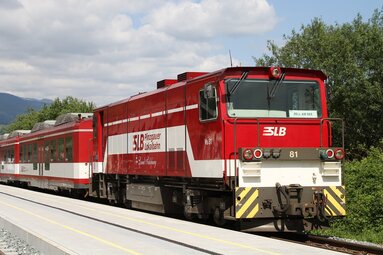 Red and white Pinzgauer Lokalbahn train at a platform, with trees and mountains in the background. | © Wikipedia
