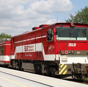 Rot-weiße Pinzgauer Lokalbahn an einem Bahnsteig, im Hintergrund Bäume und Berge. | © Wikipedia