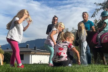 Mehrere Kinder und zwei Erwachsene stehen auf einer Wiese bei einem Pony, auf dem ein Kind mit Reithelm sitzt. | © Auszeit PFERD, Katharina Gläser